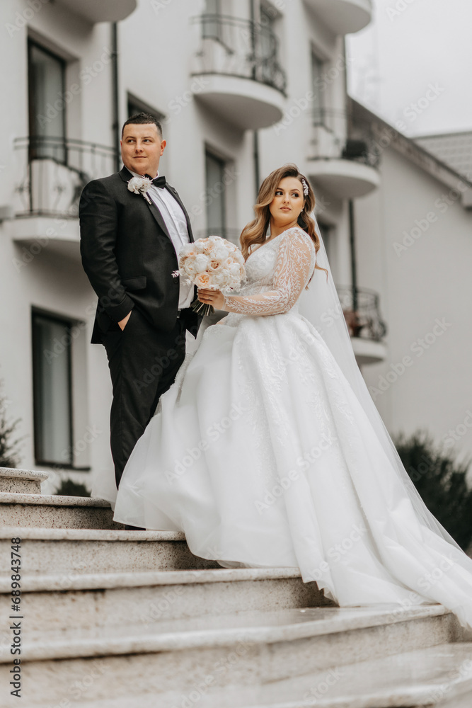 The bride and groom are walking near the hotel and posing, happy and enjoying the day, holding hands. A long train on the dress. Winter wedding