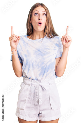 Young beautiful blonde woman wearing tye die tshirt amazed and surprised looking up and pointing with fingers and raised arms.