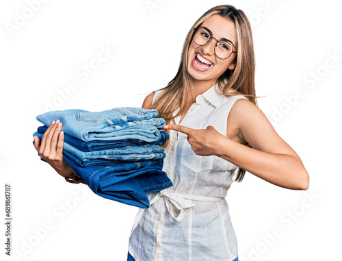 Hispanic young woman holding stack of folded jeans smiling happy pointing with hand and finger