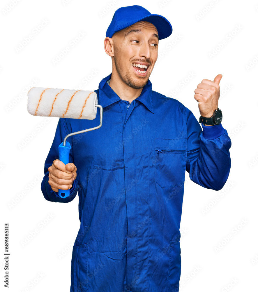 Bald man with beard holding roller painter pointing thumb up to the side smiling happy with open mouth Bald man with beard holding roller painter pointing thumb up to the side smiling happy with open mouth