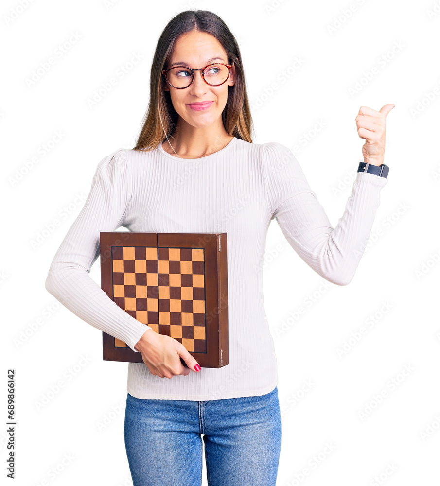 Beautiful brunette young woman holding chess pointing thumb up to the side smiling happy with open mouth Beautiful brunette young woman holding chess pointing thumb up to the side smiling happy with open mouth