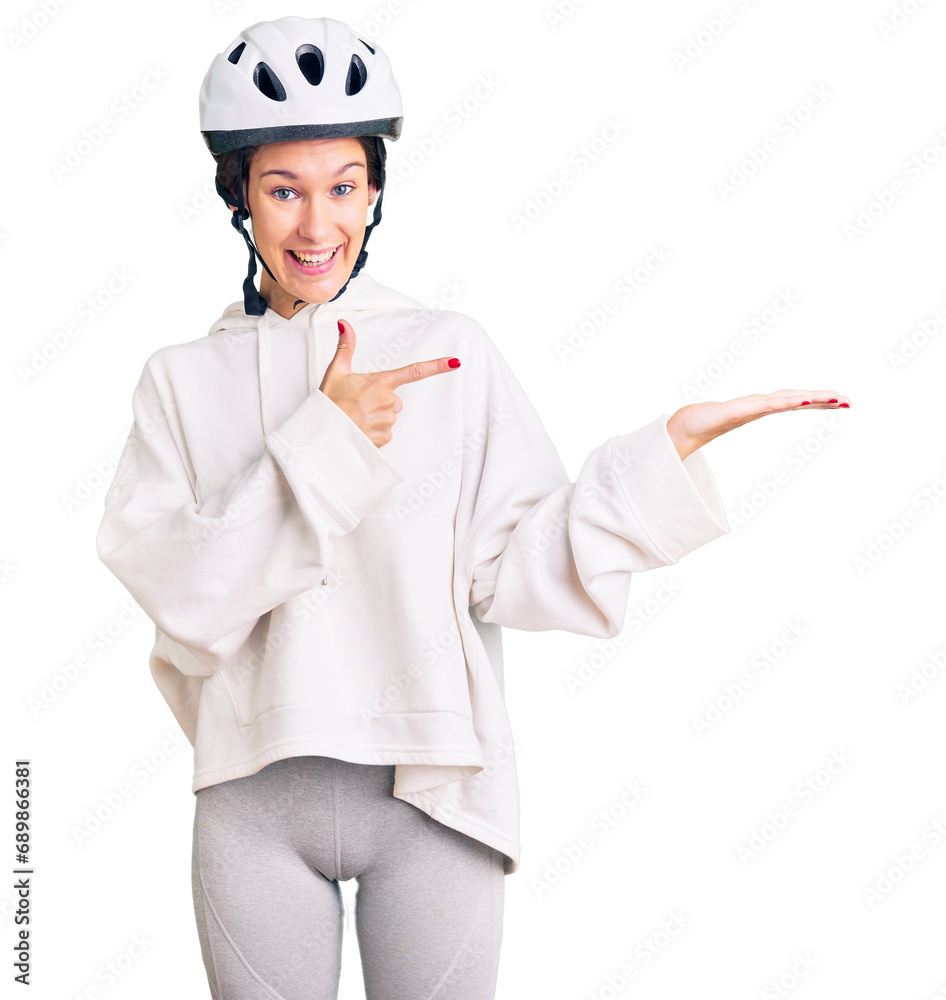 Beautiful brunette young woman wearing bike helmet and sporty clothes amazed and smiling to the camera while presenting with hand and pointing with finger. Beautiful brunette young woman wearing bike helmet and sporty clothes amazed and smiling to the camera while presenting with hand and pointing with finger.