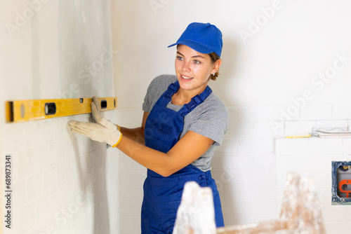 Young woman worker in the uniform uses a spirit level on the bathroom wall during renovation work in the apartment