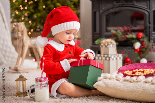 A little girl dressed as Santa Claus looks curiously into a gift box in a living room against the backdrop of a Christmas tree.
