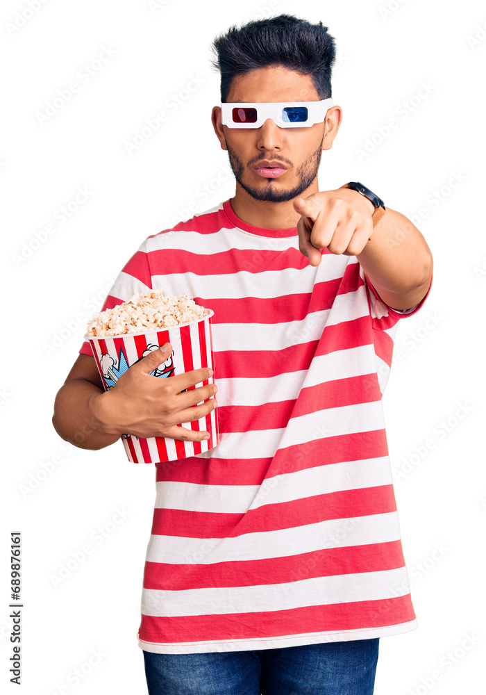 Handsome latin american young man wearing 3d glasses and eating popcorn at the movies pointing with finger to the camera and to you, confident gesture looking serious Handsome latin american young man wearing 3d glasses and eating popcorn at the movies pointing with finger to the camera and to you, confident gesture looking serious