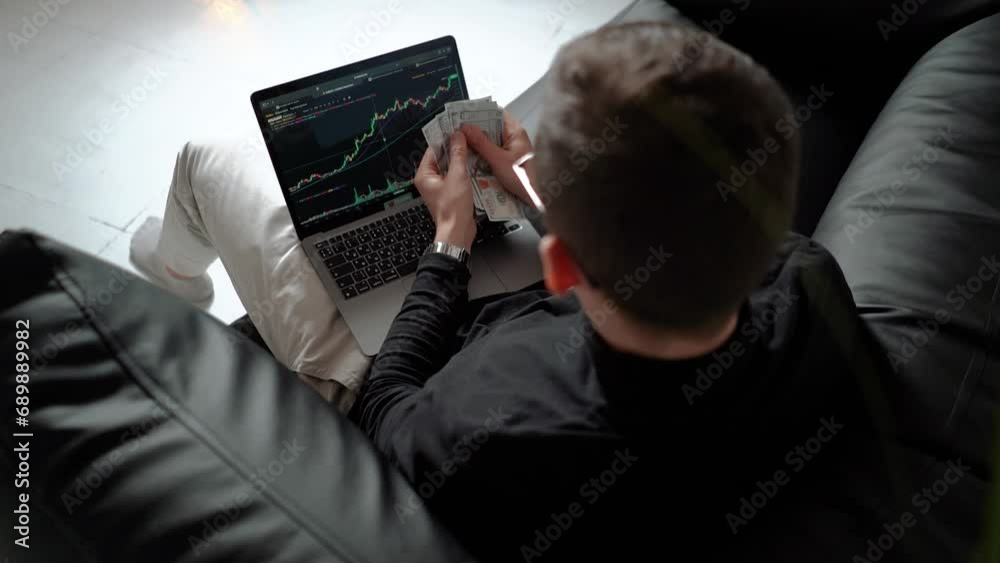 a male businessman investor trader sits with a laptop and joyfully counts his money dollars. Crypto market and trading exchange charts on the screen. Bitcoin returns and profits during the bull market