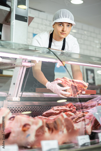 Positive young salesman standing behind counter demonstrating piece of meat in butcher shop