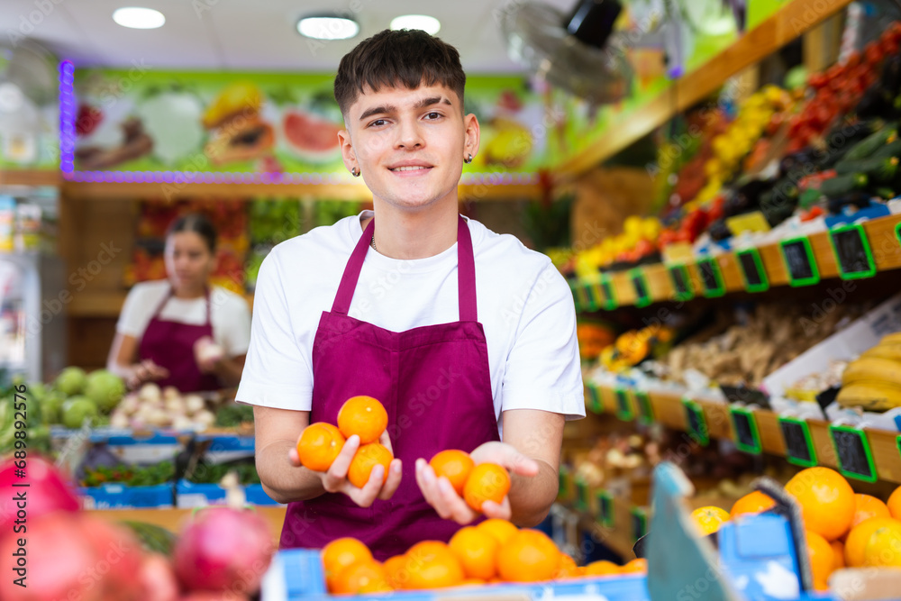 Portrait of cheerful young male in apron selling organic tangerines and fruits in supermarket