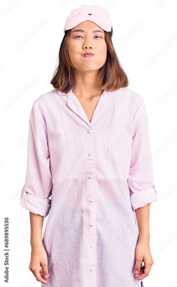 Φωτογραφία Young beautiful chinese girl wearing sleep mask and pajama puffing cheeks with funny face