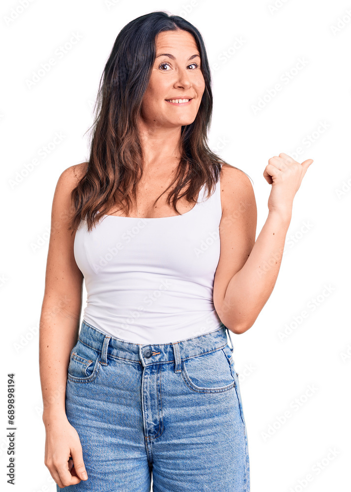 Young beautiful brunette woman wearing casual sleeveless t-shirt smiling with happy face looking and pointing to the side with thumb up. Young beautiful brunette woman wearing casual sleeveless t-shirt smiling with happy face looking and pointing to the side with thumb up.