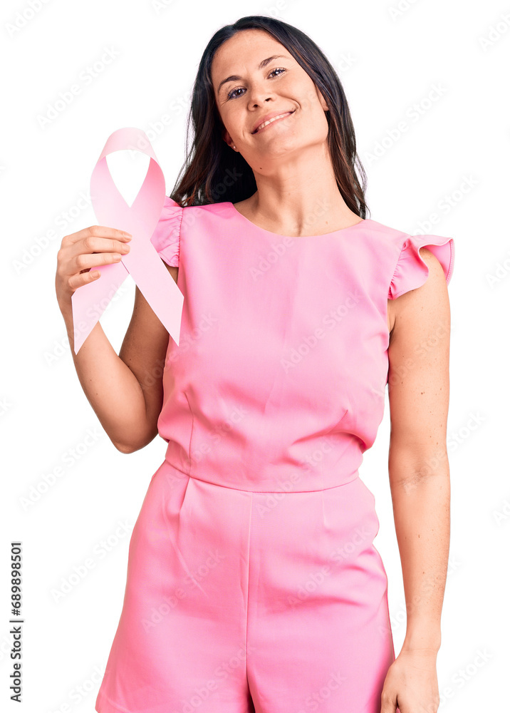Young beautiful brunette woman holding pink cancer ribbon looking positive and happy standing and smiling with a confident smile showing teeth Young beautiful brunette woman holding pink cancer ribbon looking positive and happy standing and smiling with a confident smile showing teeth