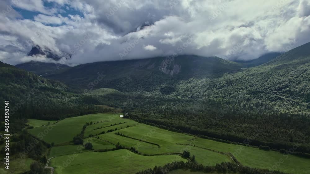 Drone rising above green meadows on the edge of vast forests, mountain ridges and mountain peak covered with white clouds in norwegian valley. Nature travel concept Drone rising above green meadows on the edge of vast forests, mountain ridges and mountain peak covered with white clouds in norwegian valley. Nature travel concept