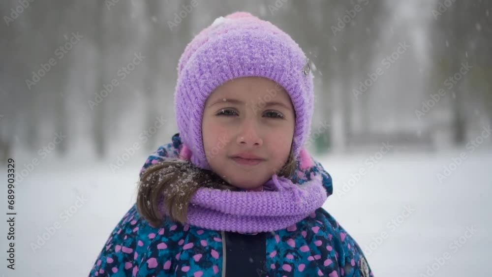 A little girl stands under snowfall in the park. A girl in a jacket and hat looks at the camera. Closeup. Slow motion. A little girl stands under snowfall in the park. A girl in a jacket and hat looks at the camera. Closeup. Slow motion.
