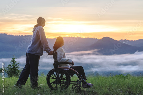 Silhouette of woman in wheelchair with care helper walking around in spring nature at sunrise misty mountains. photo