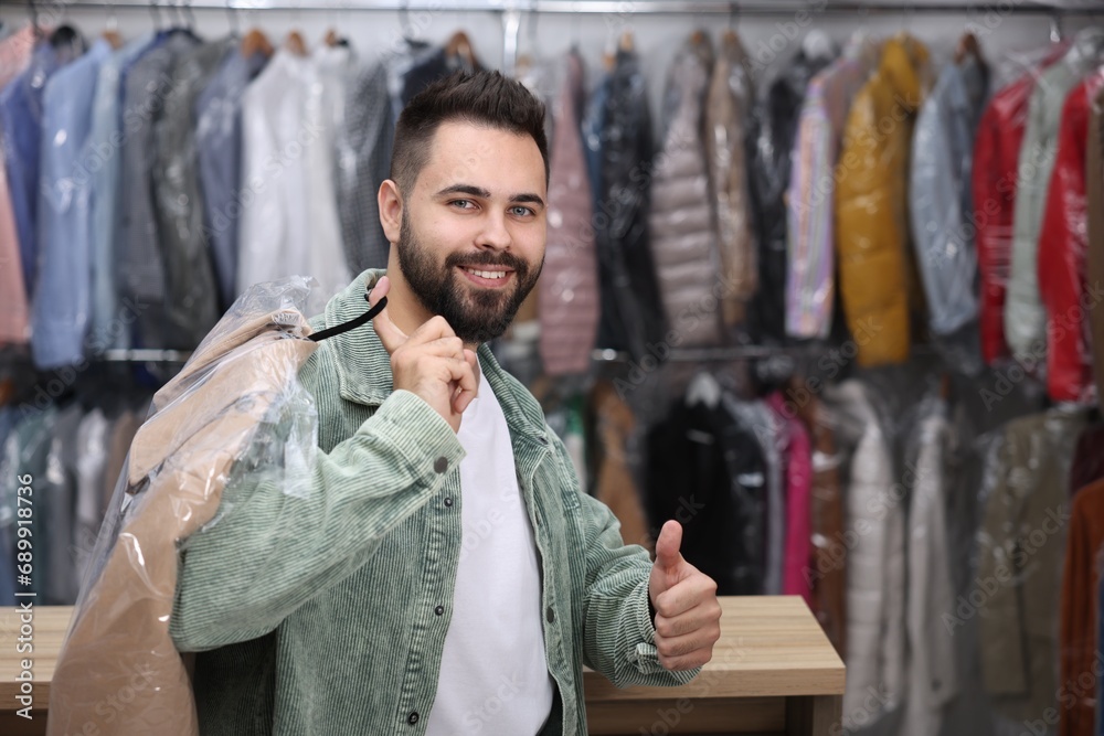 Dry-cleaning service. Happy man holding hanger with coat in plastic bag and showing thumb up indoors, space for text Dry-cleaning service. Happy man holding hanger with coat in plastic bag and showing thumb up indoors, space for text