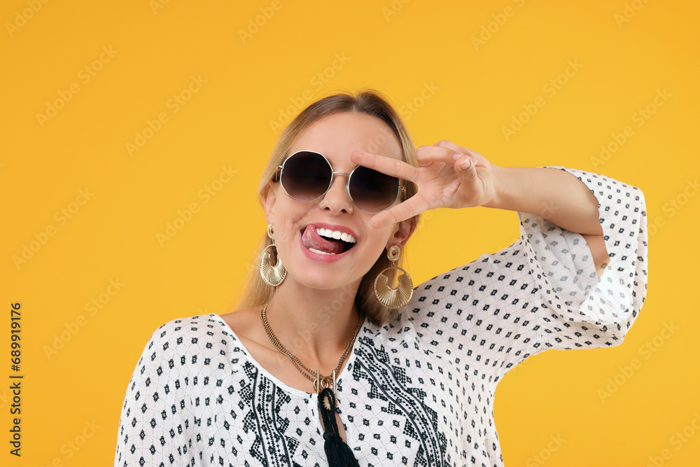 Fototapeta premium Portrait of hippie woman showing tongue and peace sign on yellow background