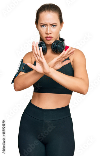 Beautiful blonde woman wearing gym clothes and using headphones rejection expression crossing arms doing negative sign, angry face