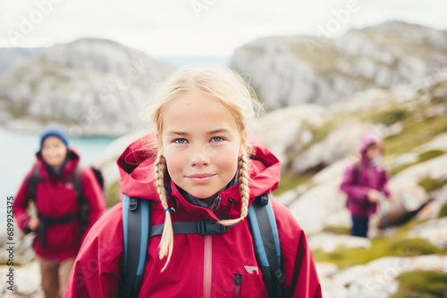 A young girl wearing red jacket with a backpack stands on top of a mountain and admires the panorama