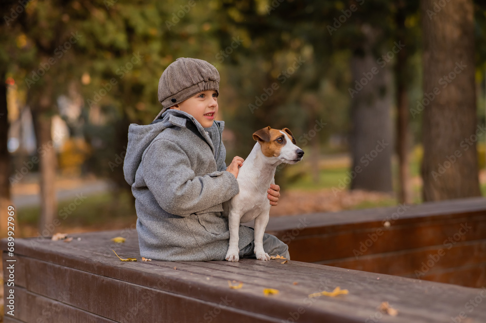 Caucasian boy sits on a bench with a dog Jack Russell Terrier for a walk in the autumn park. 