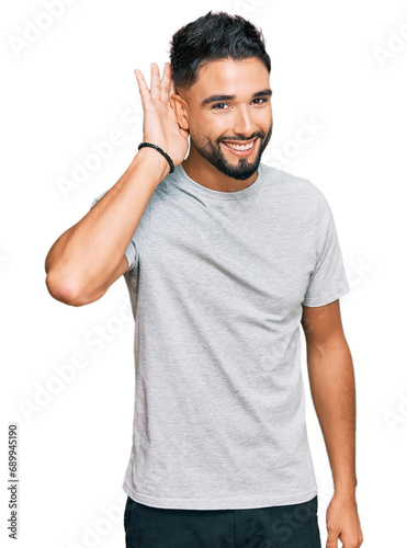 Young man with beard wearing casual grey tshirt smiling with hand over ear listening an hearing to rumor or gossip. deafness concept.