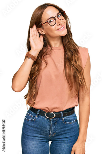 Young hispanic woman wearing casual clothes and glasses smiling with hand over ear listening an hearing to rumor or gossip. deafness concept.
