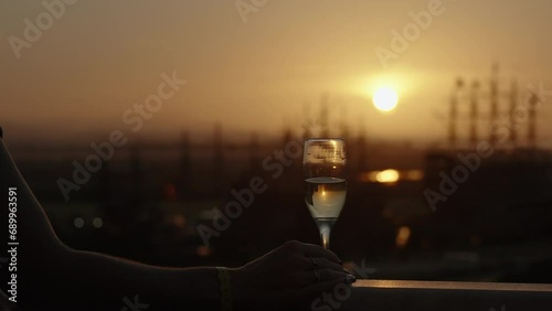 A woman steps onto the balcony to bid farewell to the sunset with a glass of champagne, tall masts of sailing ships in the distance at sea. photo
