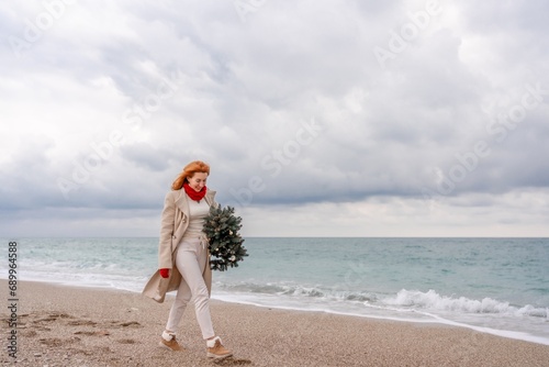 Redhead woman Christmas tree sea. Christmas portrait of a happy redhead woman walking along the beach and holding a Christmas tree in her hands. Dressed in a light coat, white suit and red mittens.