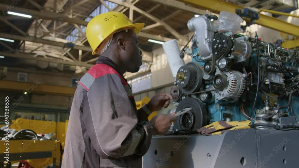 Side medium shot of African American male technician in yellow hardhat and overalls inspecting pulleys of large explosion engine and then typing information into tablet at machinery factory Side medium shot of African American male technician in yellow hardhat and overalls inspecting pulleys of large explosion engine and then typing information into tablet at machinery factory