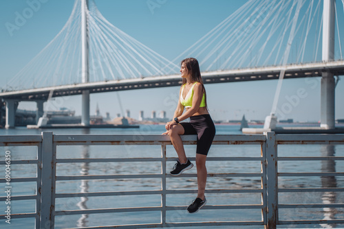 Fit caucasian young woman in sportswear, sneakers sitting on fence at embankment looks forward on bay against ocean bay and bridge. Sport, active people. Youth.