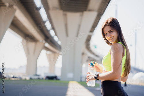 Cheerful young woman in sporty leggings and yellow top holds bottle of water relaxing after jogging outdoors looks at camera broad smiling. Successful sport person.