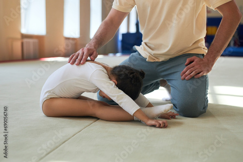 Brunette girl in white gymnastic clothing stretching bending over her legs while coach holding hand on her back, copy space