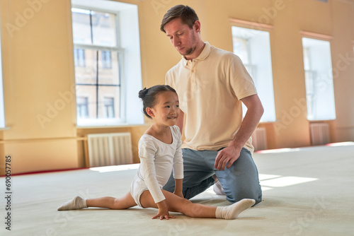 Wide shot of smiling Asian little girl sitting in front split while focused Caucasian male gymnastics instructor observing her technique