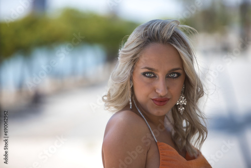 Portrait of beautiful young woman, blonde with blue eyes and an orange dress, leaning on a railing looking at camera with her beautiful eyes. Concept beauty, fashion, looks, eyes.