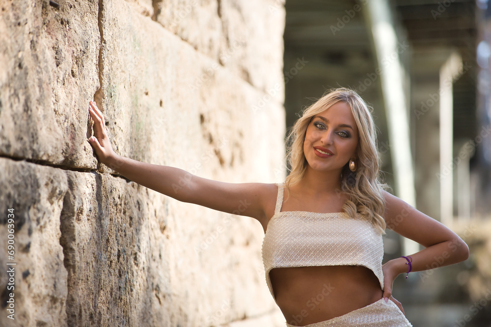 Young and beautiful woman, blonde with blue eyes and wearing a white sequined dress, leaning against a stone wall, looking tender and dreamy. Concept beauty, fashion, trend, dreams, tenderness.
