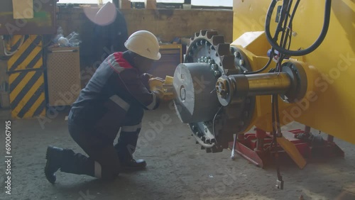Side of workman in hardhat and overall bolting up large gear on tractor framework using wrench at heavy machinery production factory on sunny day