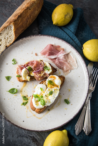 Italienische Burrata Käse Crostini aus Ciabatta Brot,konfierter Knoblauch, Prosciutto Schinken, rote Zwiebel, Basilikum, Honig, Olivenöl, Zitronen mit Teller, Besteck, Serviette blau, dunkel grau Stei