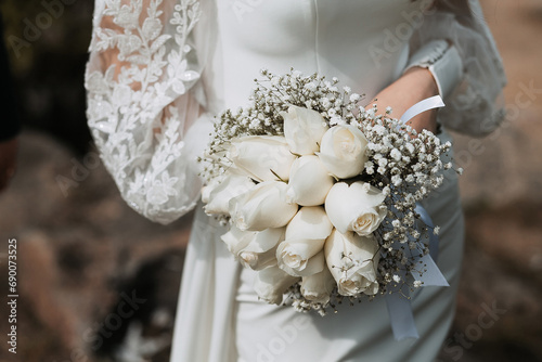 The bride in a long tight white wedding dress holds a beautiful bouquet of white roses in her hands during the day. Image for your design or creative compositions.