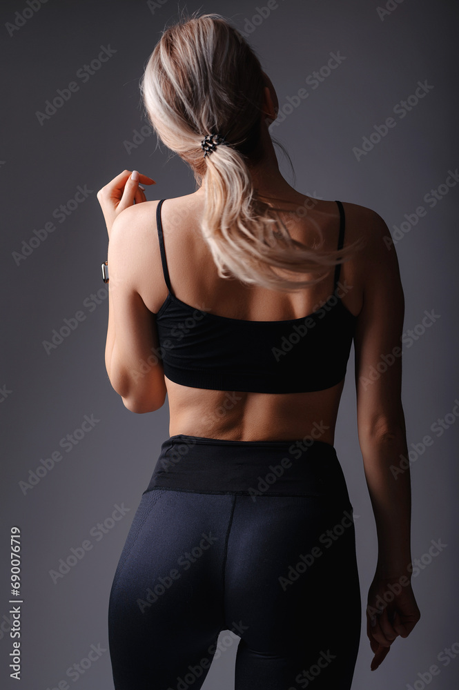 Sporty young woman in sportswear showing her figure on grey studio background. Back view of blonde fitness girl wearing black top and leggings Sporty young woman in sportswear showing her figure on grey studio background. Back view of blonde fitness girl wearing black top and leggings