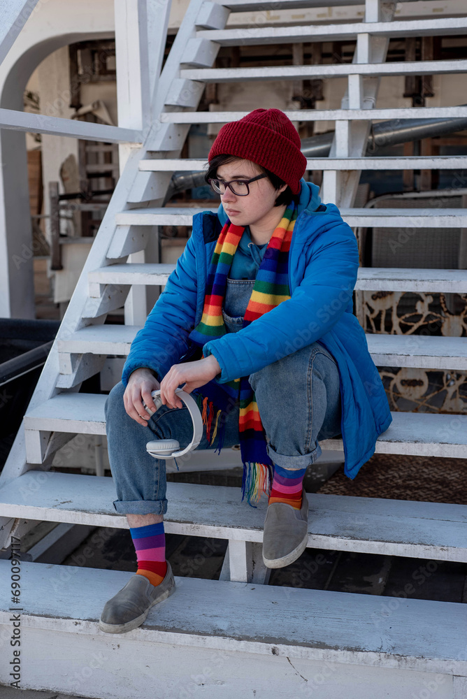Portrait of a transgender teenager in a rainbow scarf and socks, holding headphones and sitting on the stairs outdoors Portrait of a transgender teenager in a rainbow scarf and socks, holding headphones and sitting on the stairs outdoors