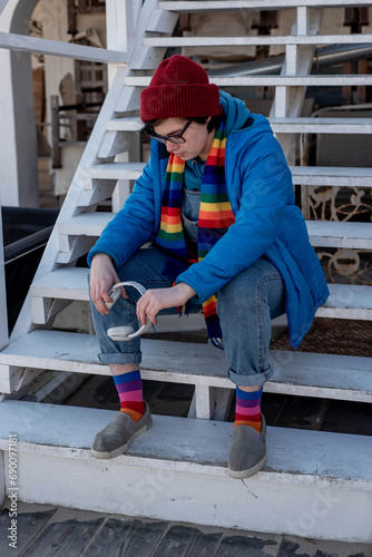 Sad transgender teenager in a rainbow scarf and socks, holding headphones and sitting on the stairs outdoors