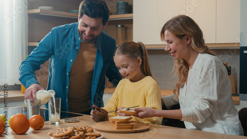 Caucasian family cooking breakfast together at kitchen at home mother with little daughter child girl prepare toasts bread with chocolate pasta father pour milk into glasses parents with kid cook