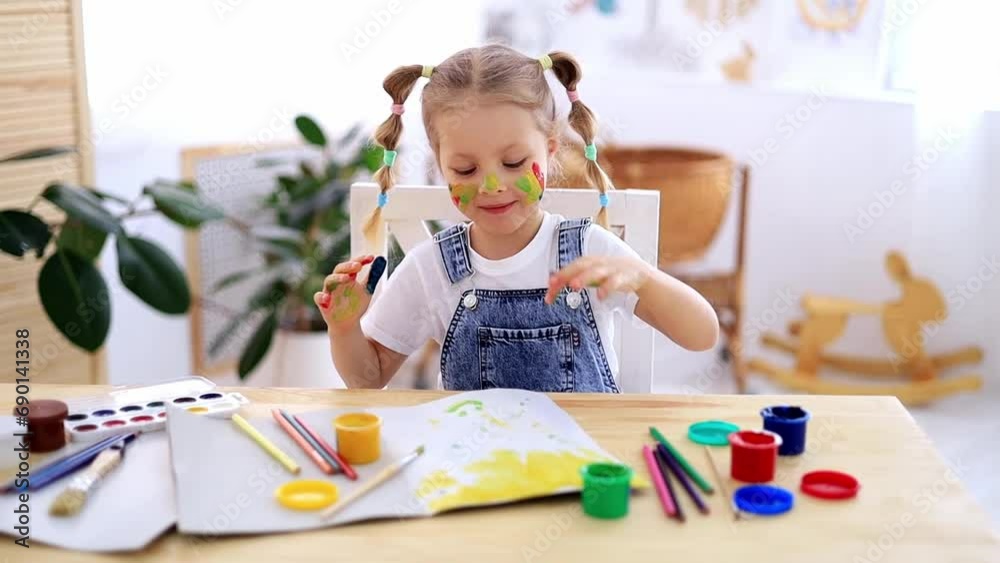 a little child girl happily makes prints in an album with her hands with a dirty face and smiles, a happy child draws a picture with gouache fingers at home at the table