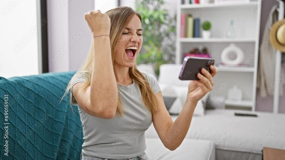 Exuberant young blonde woman, in her comfy apartment, cheering while comfortably sitting on her sofa, engrossed in playing an exciting online video game on her mobile gadget Exuberant young blonde woman, in her comfy apartment, cheering while comfortably sitting on her sofa, engrossed in playing an exciting online video game on her mobile gadget