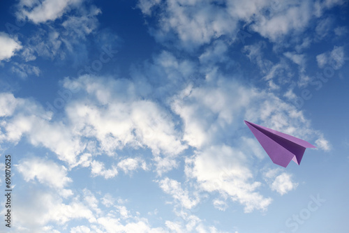 Purple paper plane flying in blue sky with clouds