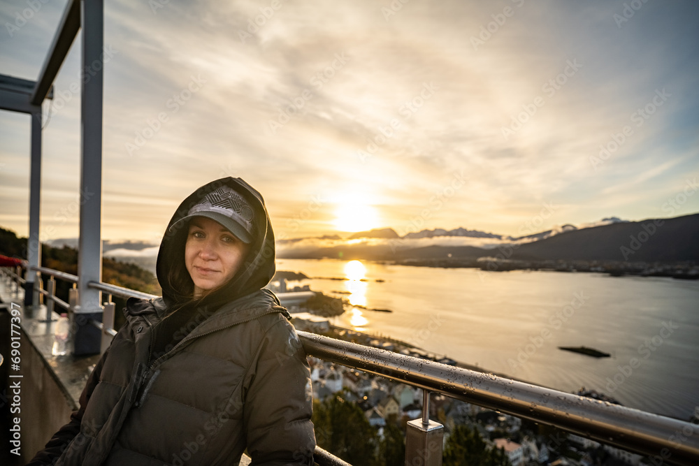 young beautiful woman in cap looking at camera with mountains on background during sunrise in alesund, norway