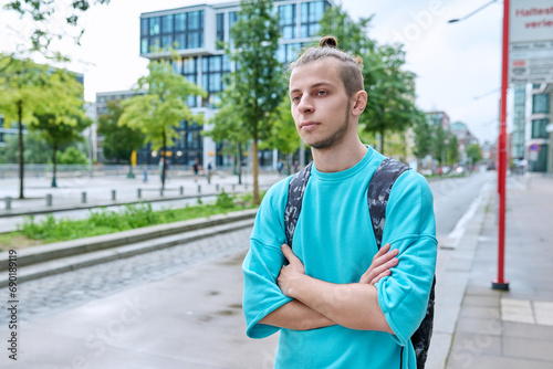 Serious young guy with backpack with crossed arms outdoor, city background