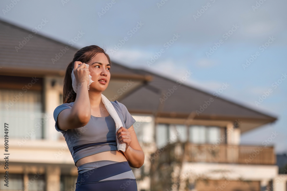 Fototapeta premium Tired and sweaty Asian woman in sportswear uses a towel to wipe away sweat. and take a break from running cardio at the park.