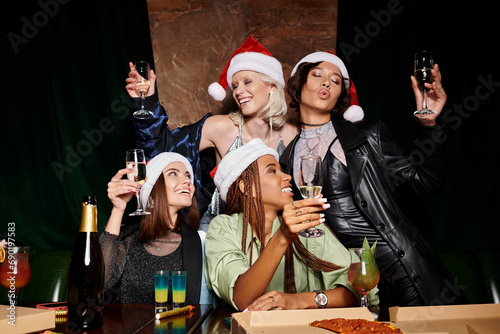 happy and trendy multiracial women in Christmas hats holding champagne and having fun in bar