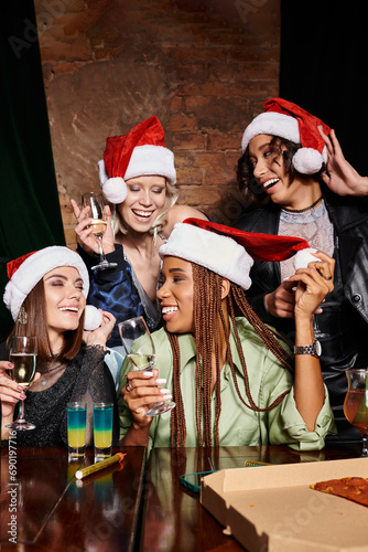 young and carefree multiethnic girlfriends in Christmas hats with champagne having fun in bar, party