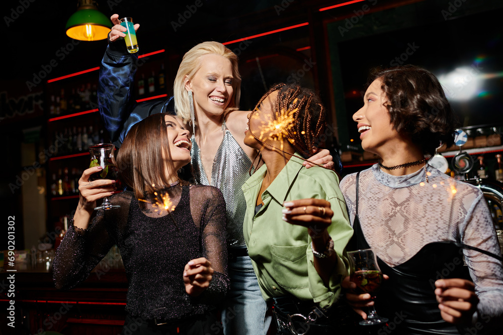 happy and stylish multiethnic girlfriends with cocktails and festive sparklers during party in bar happy and stylish multiethnic girlfriends with cocktails and festive sparklers during party in bar